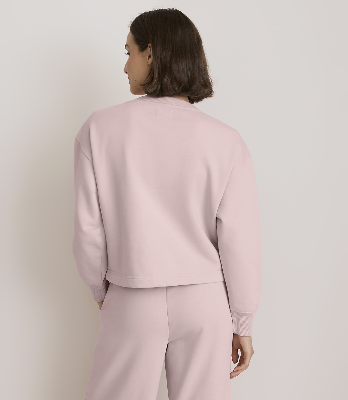 A woman in a pale pink sweatshirt and pants stands against a plain, light-colored background.
