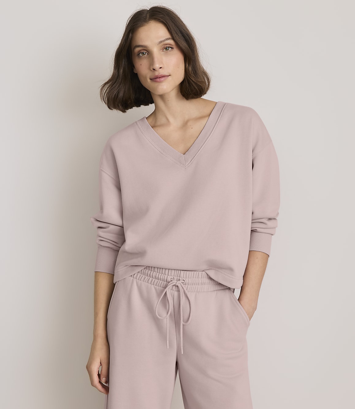 A woman in a mauve v-neck sweatshirt and matching drawstring pants stands against a plain, light gray background.