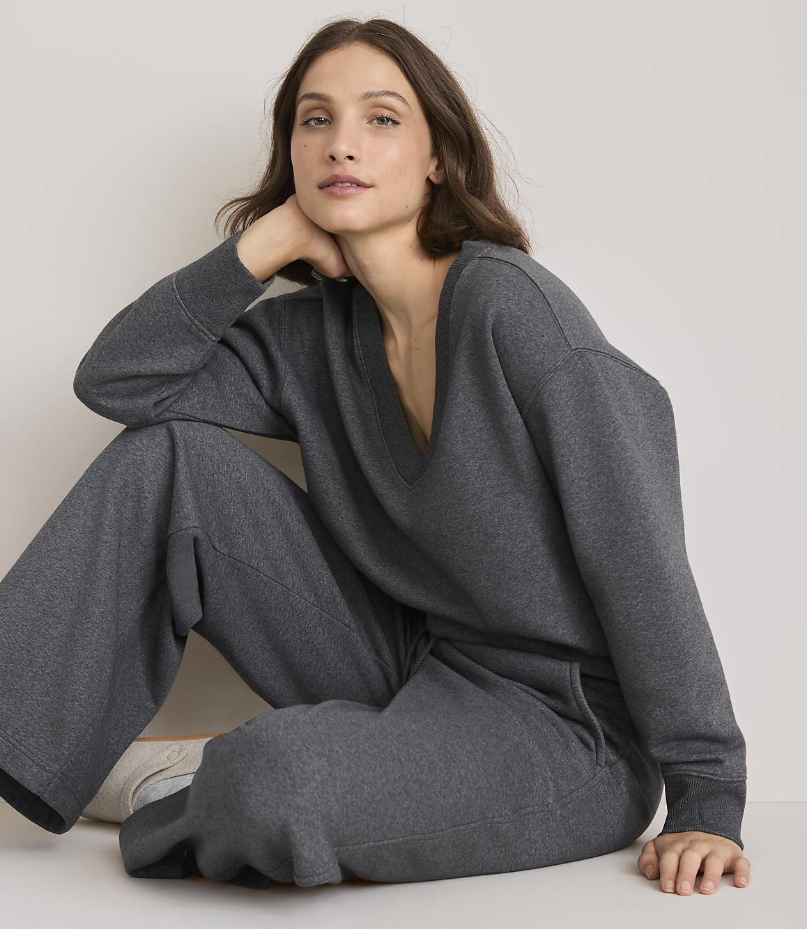 A woman in a grey loungewear set sits on a light-colored floor with a plain background.