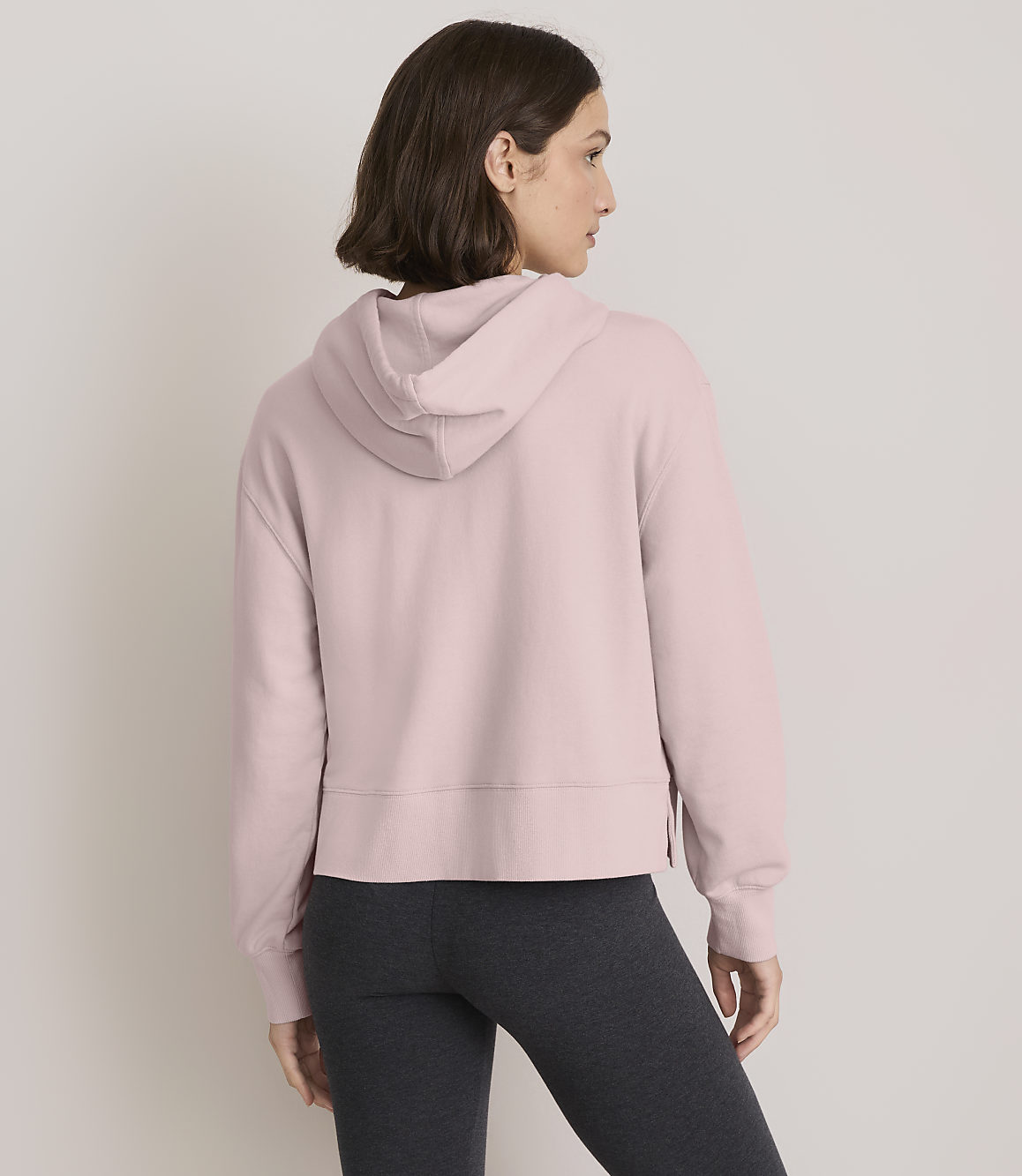 A woman in a pink hoodie and dark leggings stands against a plain, light gray background.
