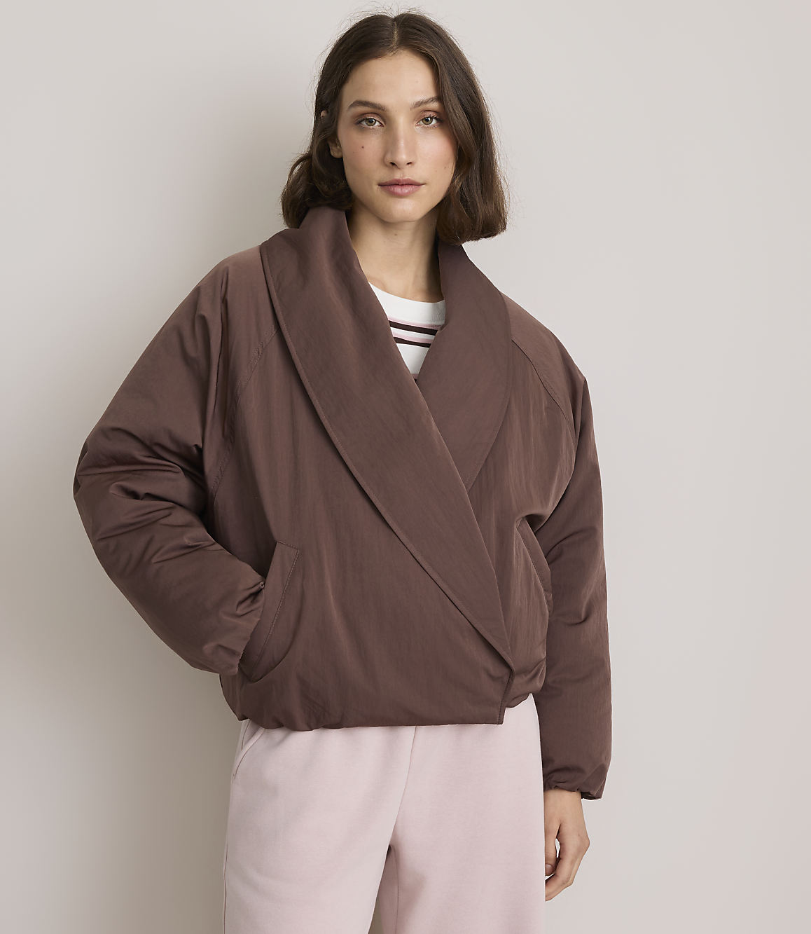 A woman in a brown puffer jacket and pink pants stands against a plain, light-colored background.