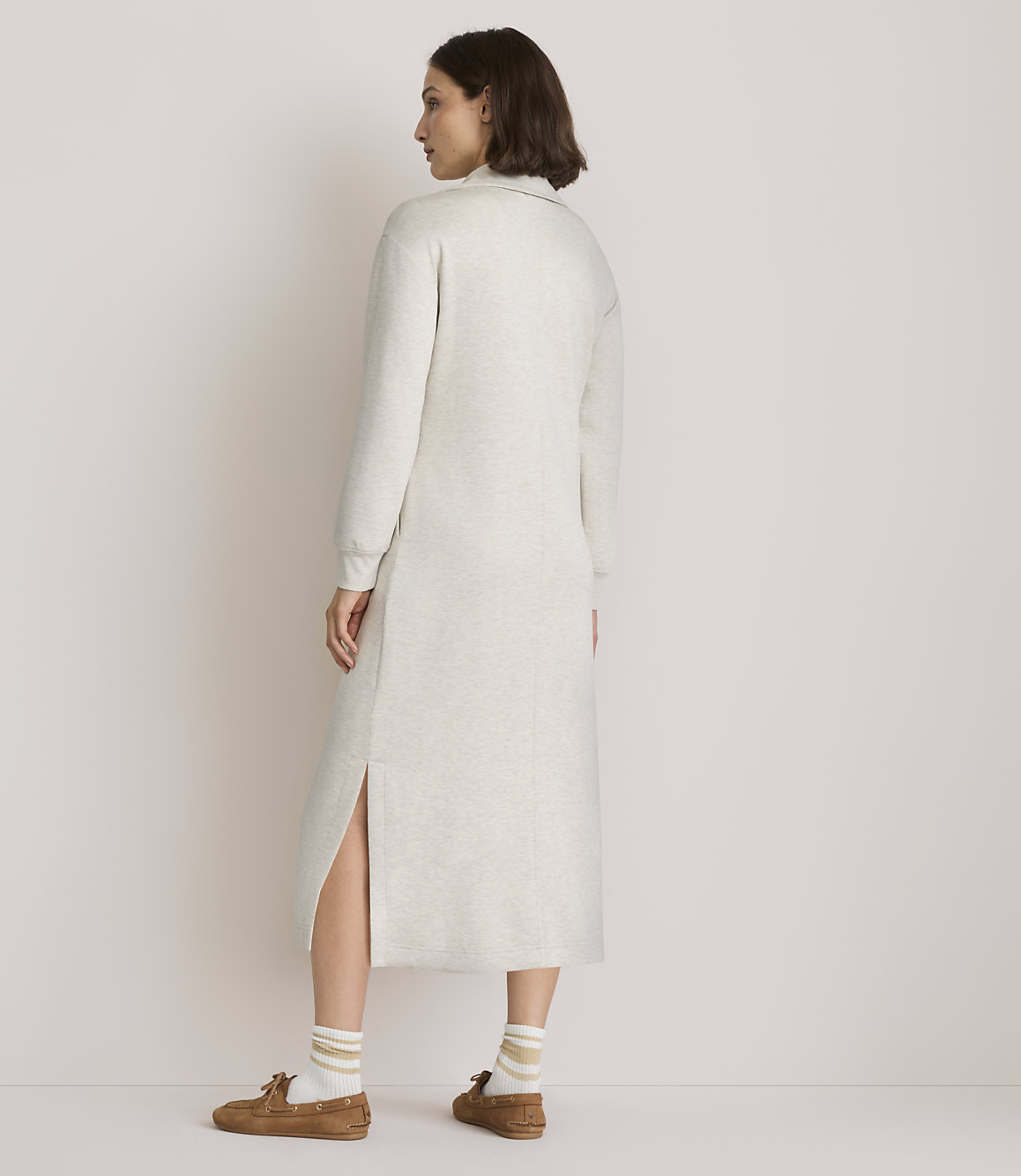 A woman in a light-colored sweater dress and loafers stands against a plain, light-colored background.