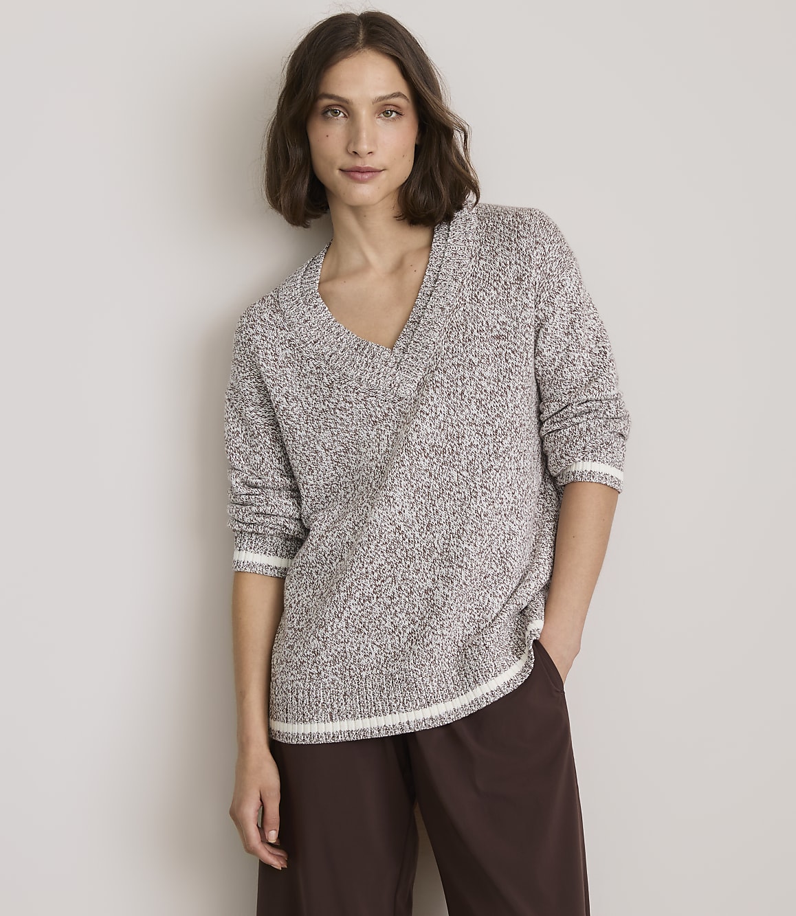A woman in a speckled v-neck sweater and brown pants stands against a plain, light gray background.