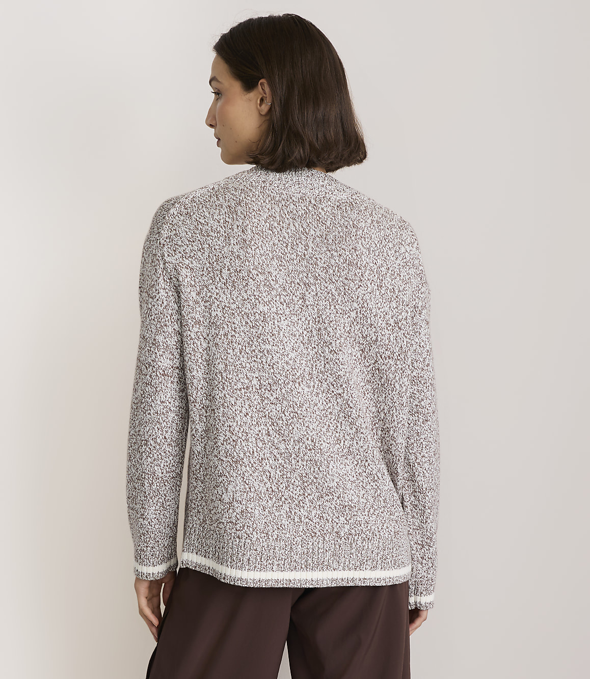 A woman in a speckled brown and white sweater stands against a plain, light brown background.