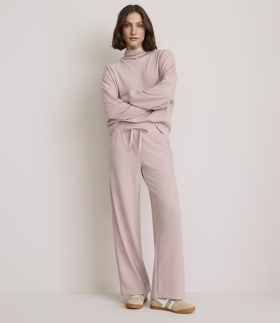 A woman in a pink ribbed loungewear set stands with her arms crossed against a plain, light-colored background.