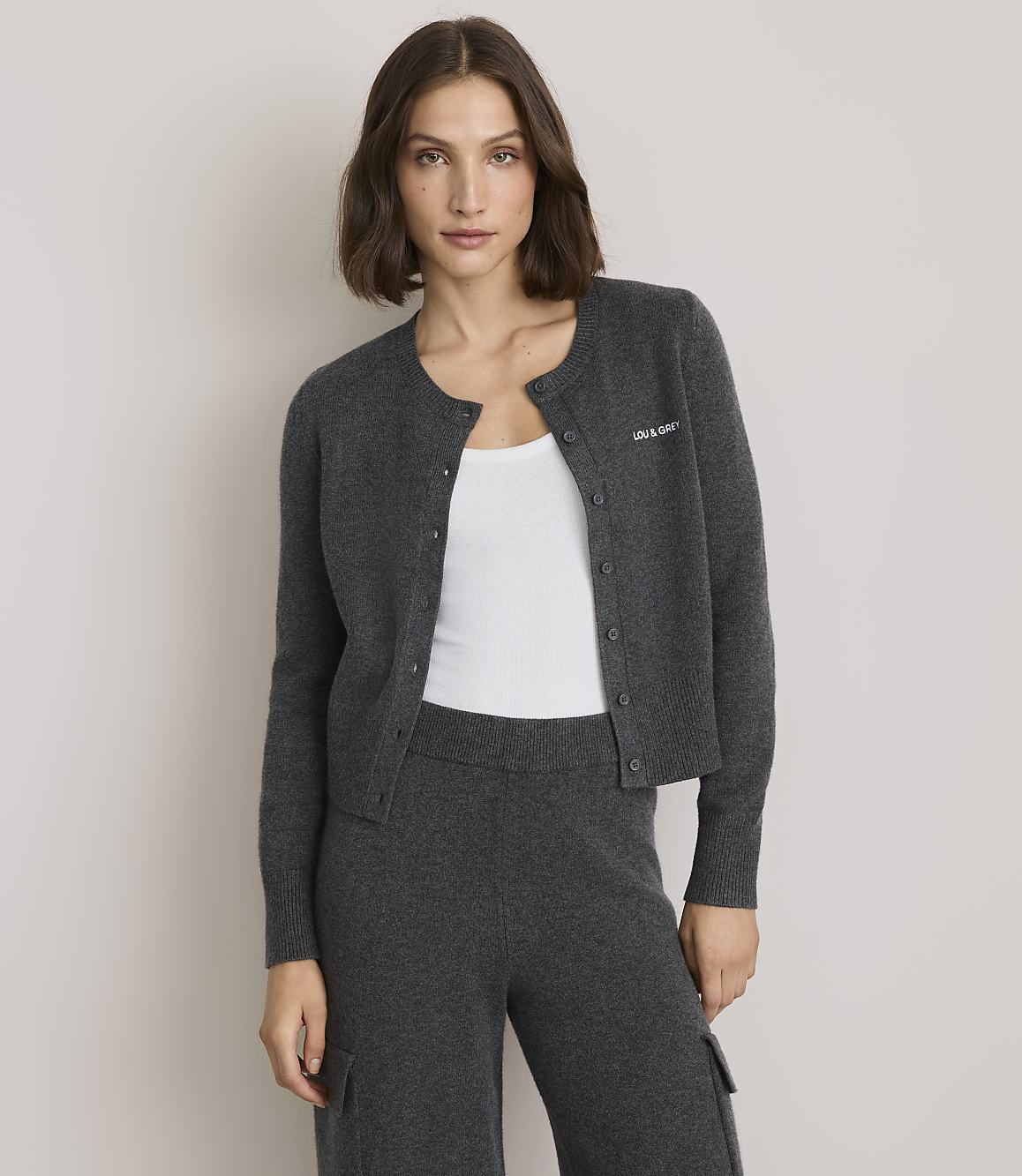 A woman in a dark gray cardigan and matching pants stands against a plain gray background.