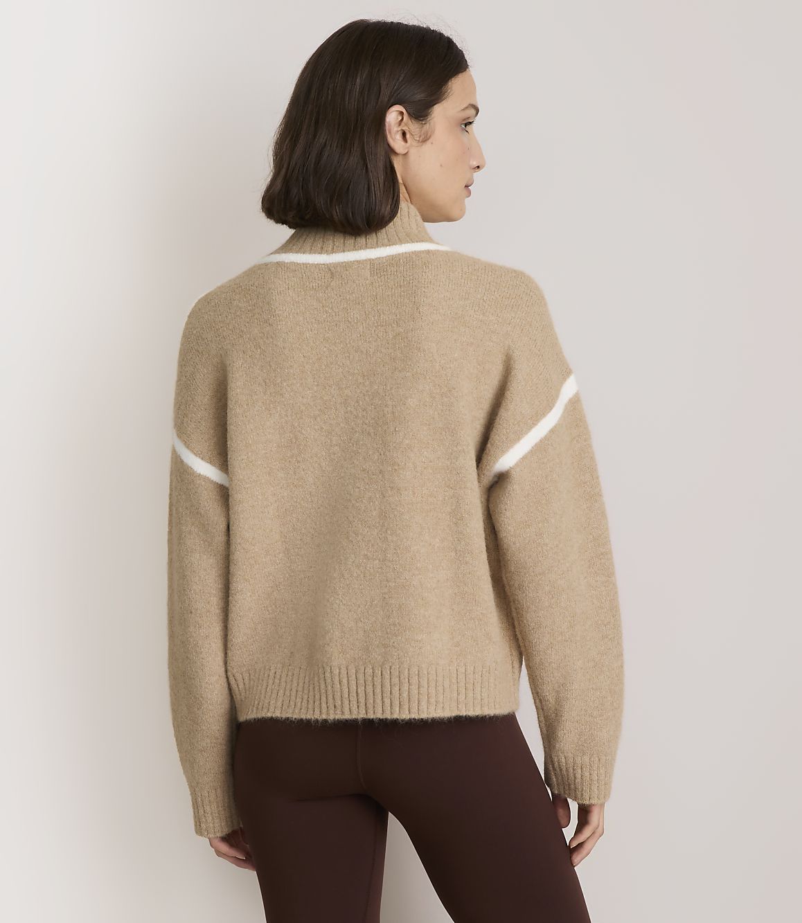 A woman in a tan sweater with white stripes stands against a plain, light-colored background.