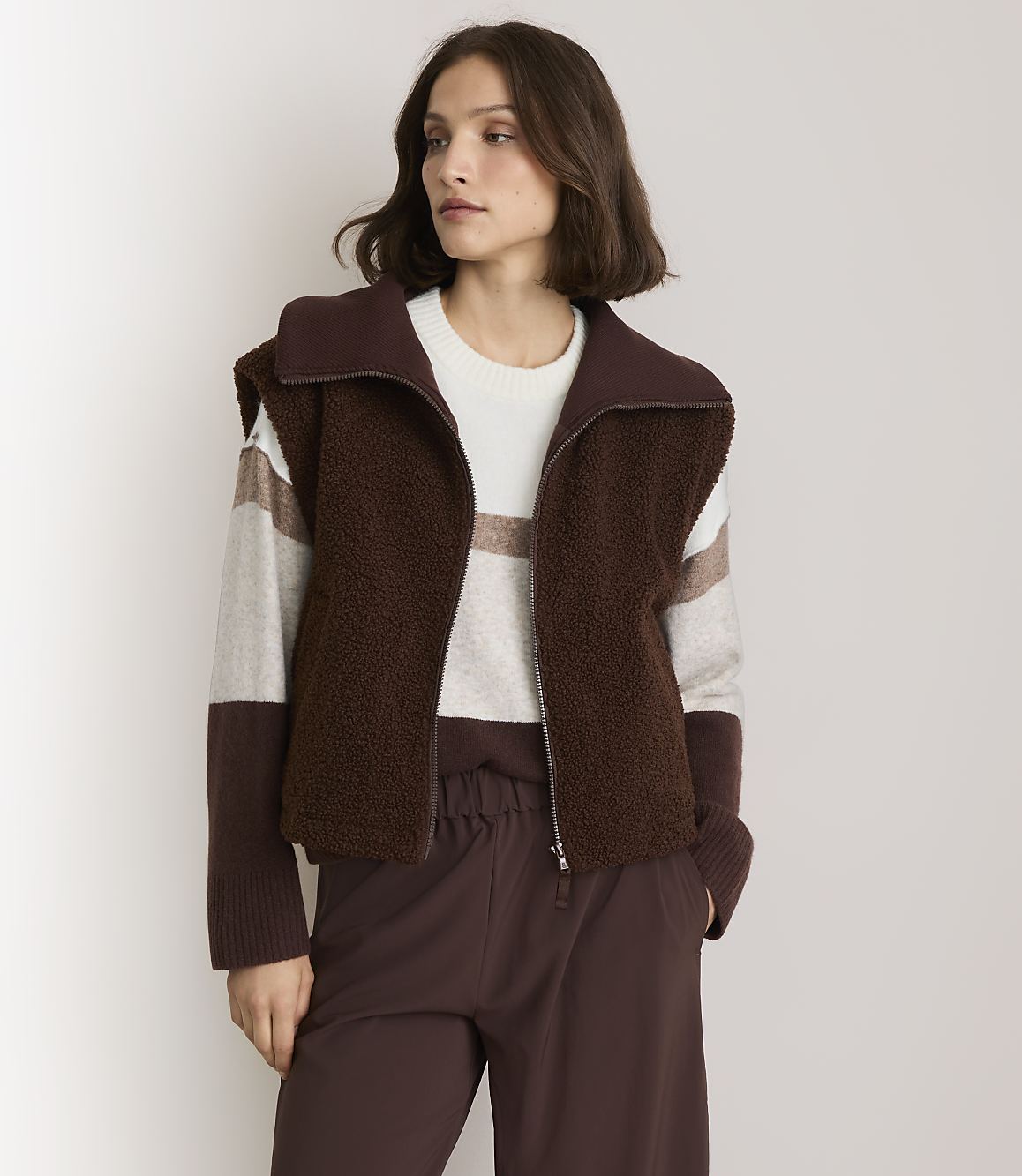 A woman in a brown fuzzy vest and striped sweater stands against a plain white background.