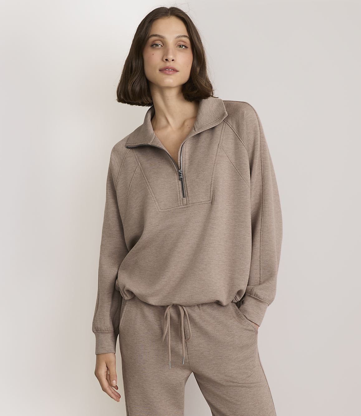 A woman in a taupe quarter-zip sweatshirt and matching sweatpants stands against a plain, light gray background.