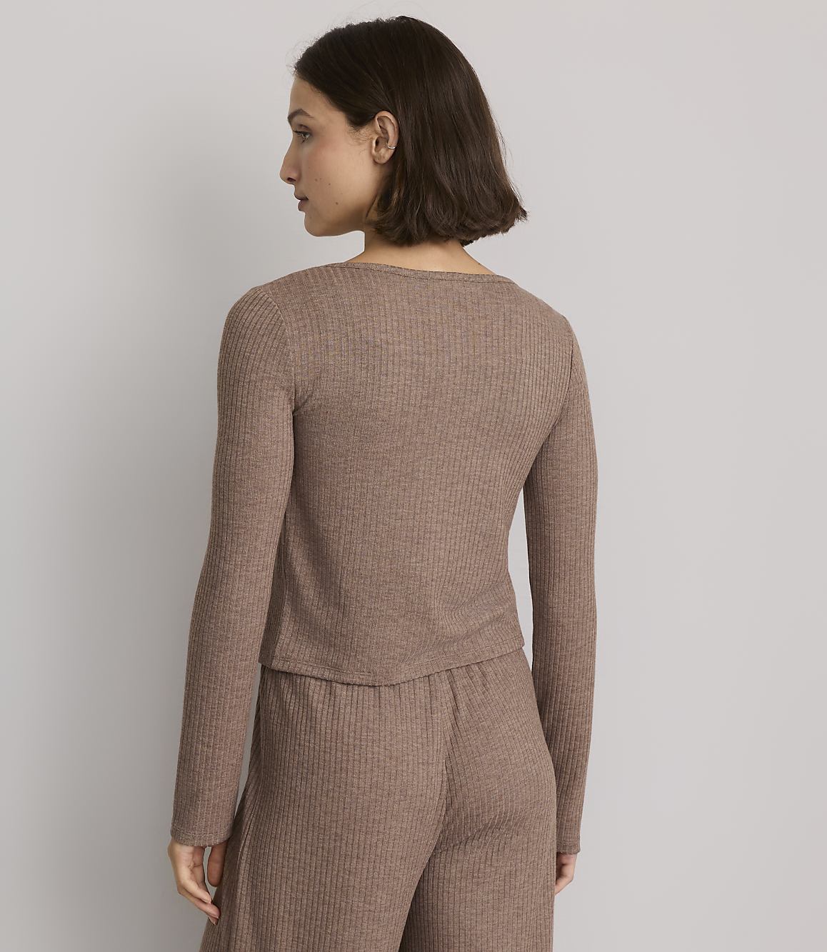 A woman in a brown ribbed top and pants stands against a plain gray background.