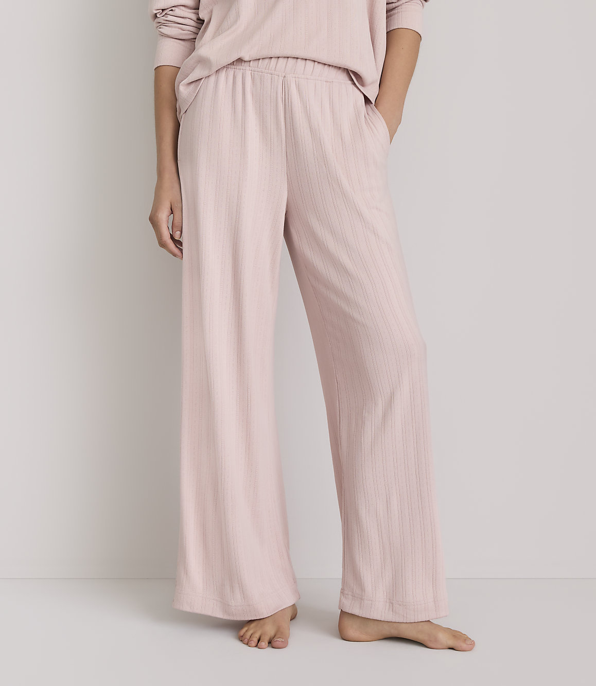 A person stands in pale pink ribbed pajama pants with their hands in their pockets against a plain white background.
