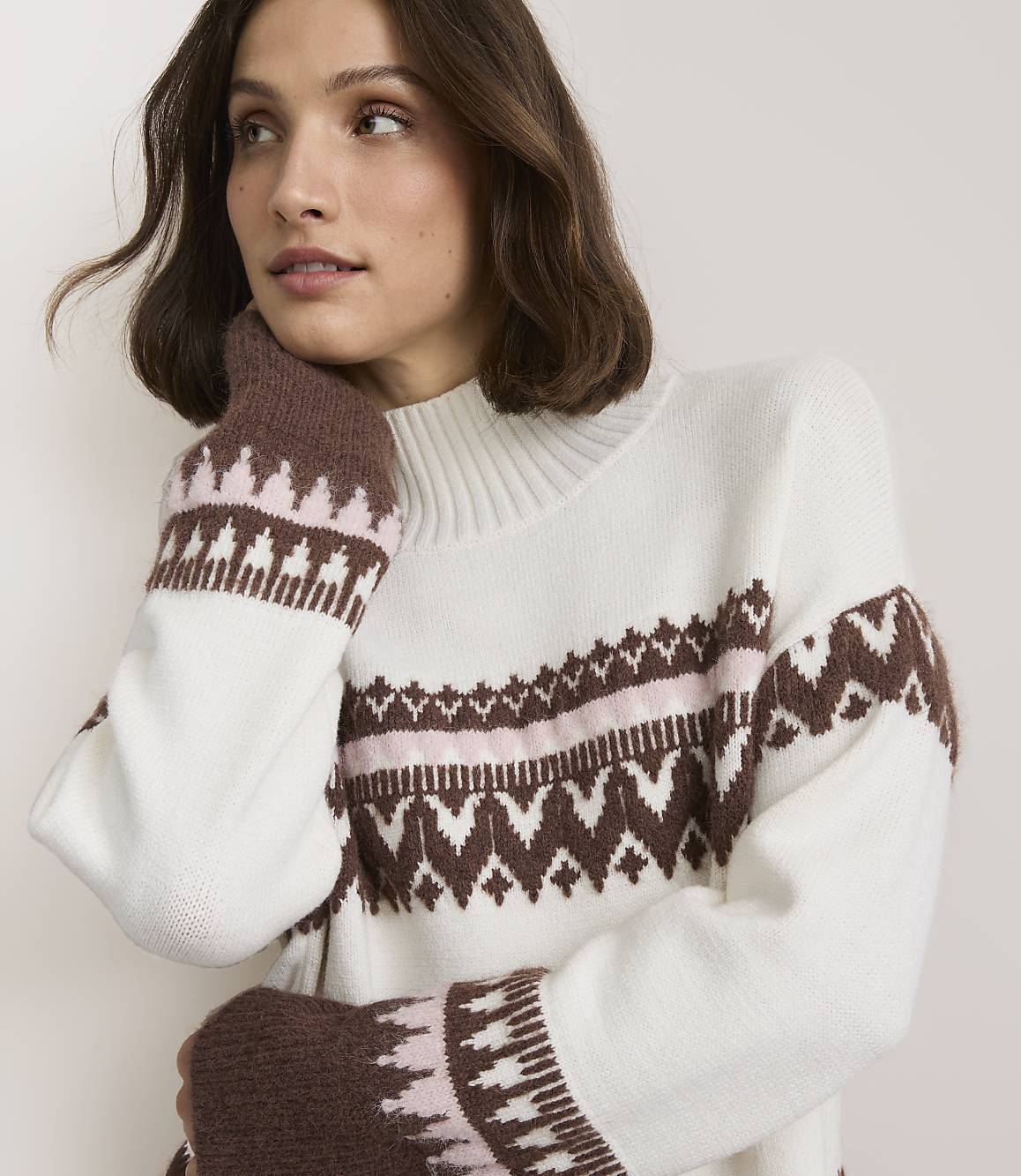 A woman in a white sweater with brown and pink geometric patterns poses against a plain white background.