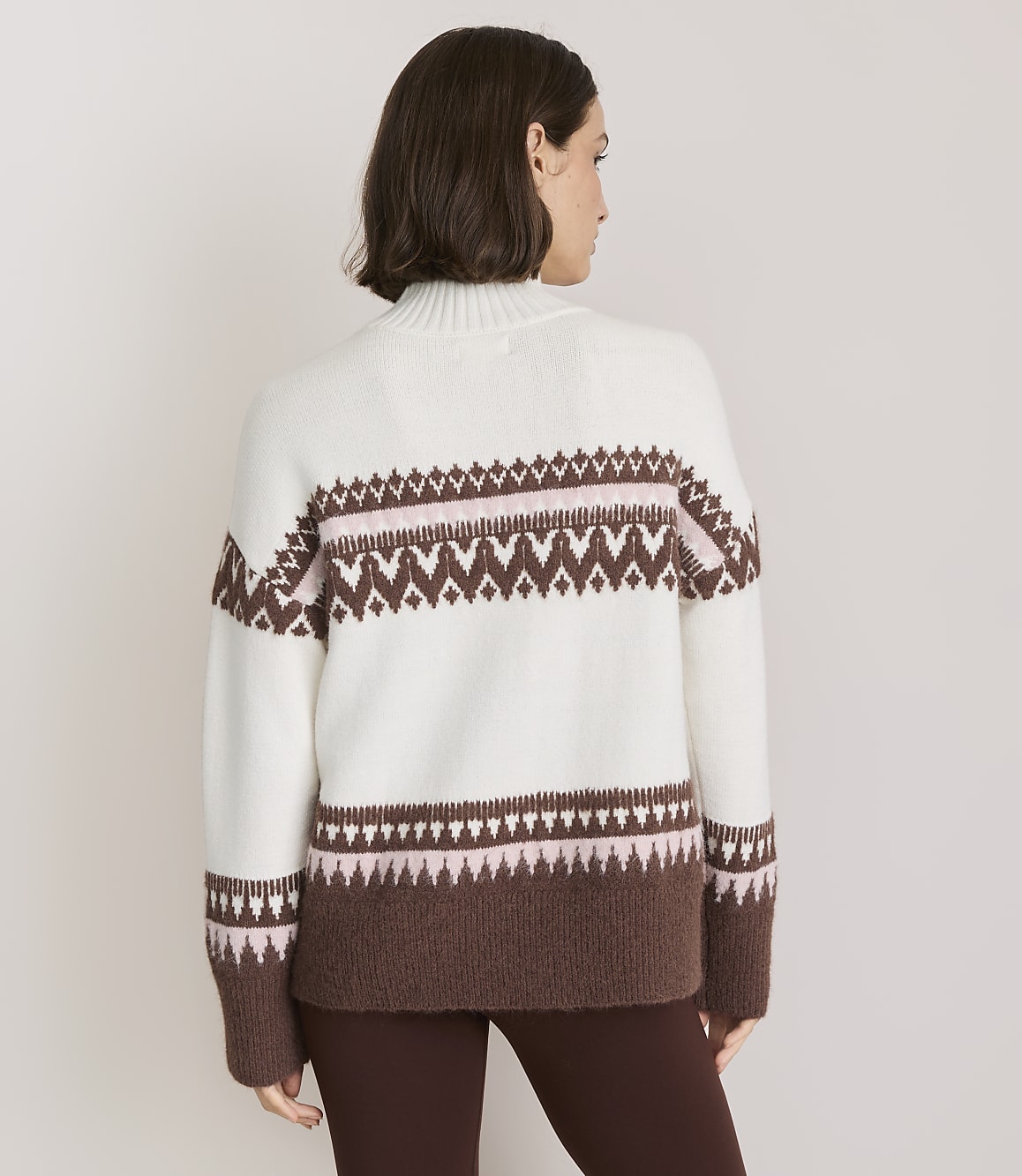A woman in a white and brown patterned sweater stands against a plain white background.