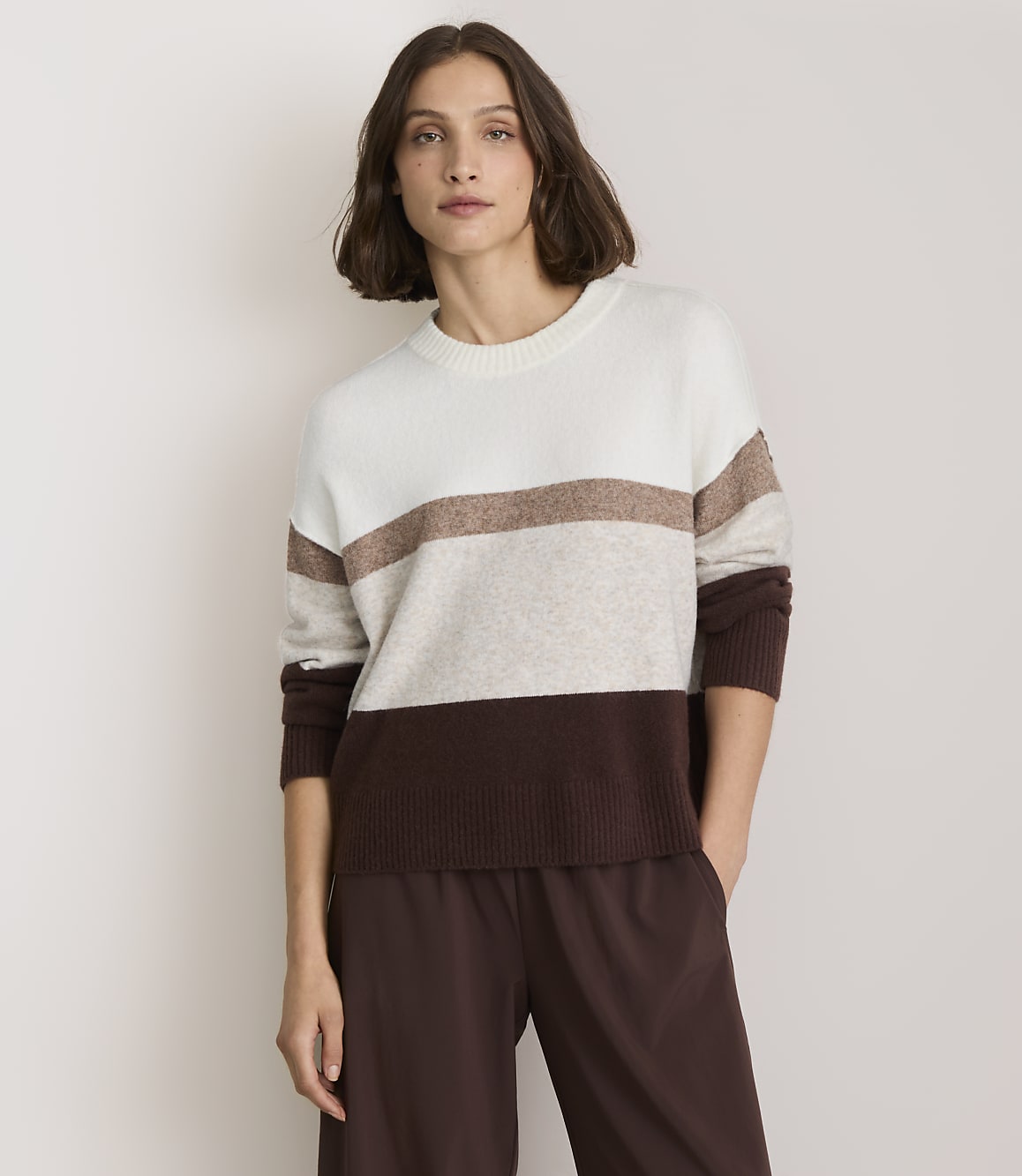 A woman in a striped sweater and brown pants stands against a plain white background.