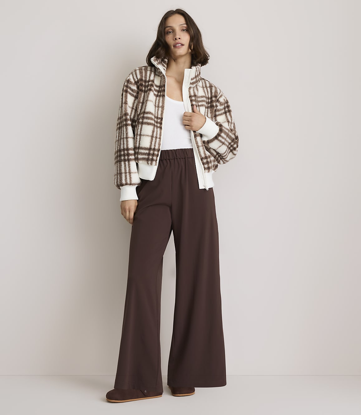 A woman in a plaid bomber jacket and wide-leg brown pants stands against a plain, light-colored background.