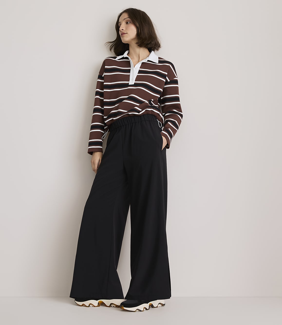 A woman in a striped rugby shirt and wide-leg black pants stands against a plain, light-colored wall.