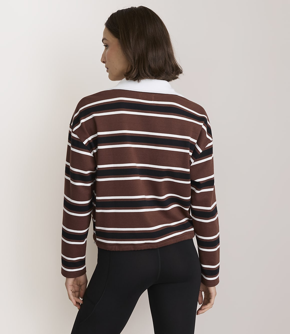 A woman in a brown, white, and black striped long-sleeved shirt and black leggings stands against a plain, light-colored background.