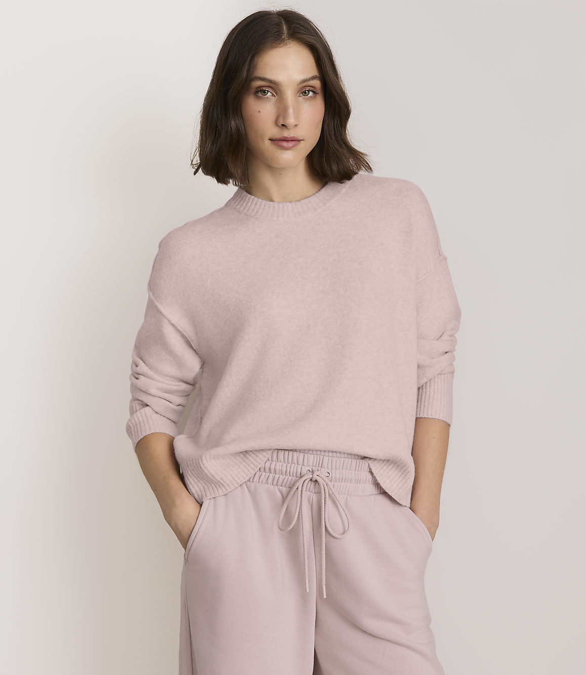 A woman in a pink sweater and matching pants stands against a plain, light background.