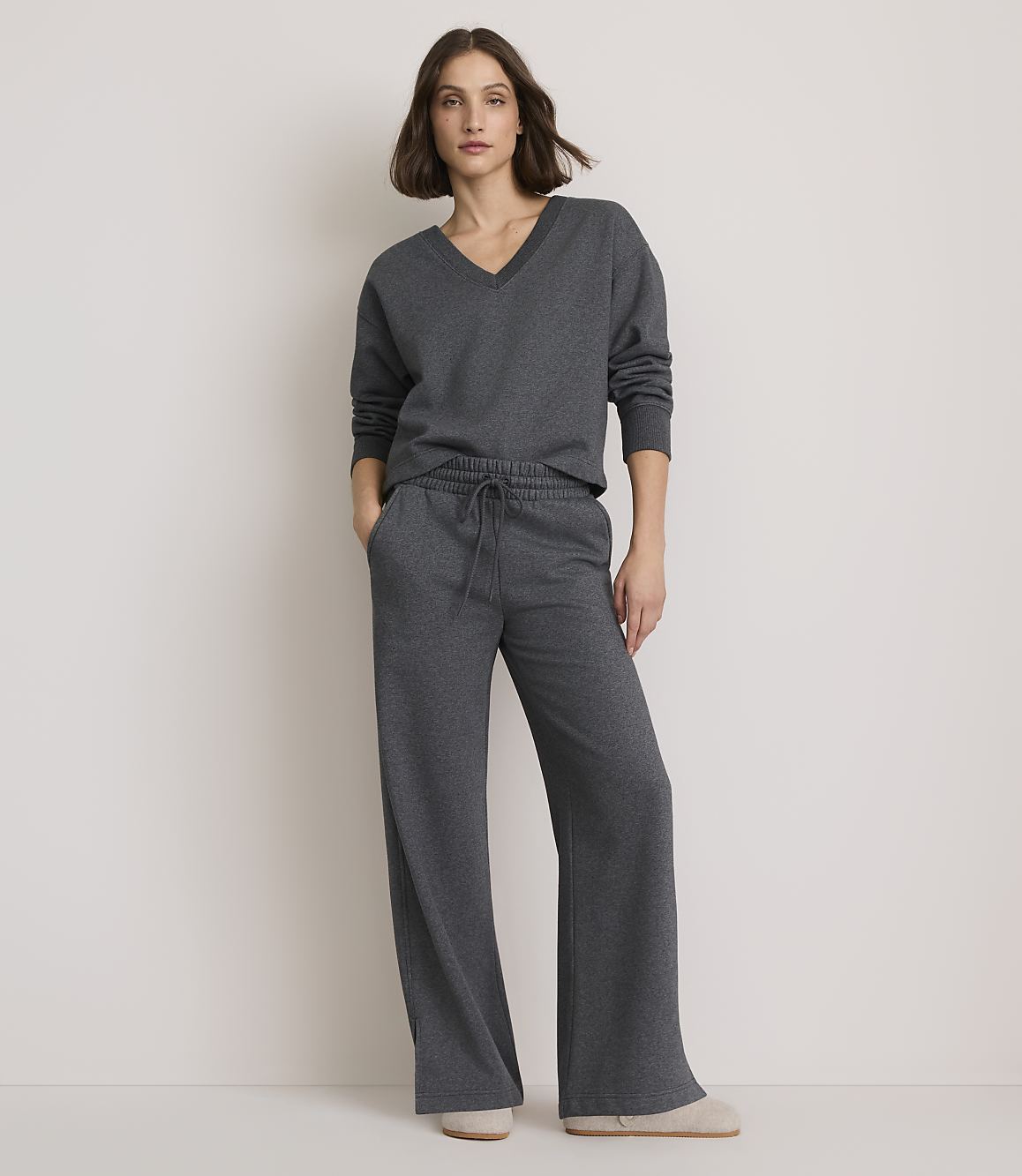 A woman in a grey v-neck sweater and wide-leg sweatpants stands against a plain white background.