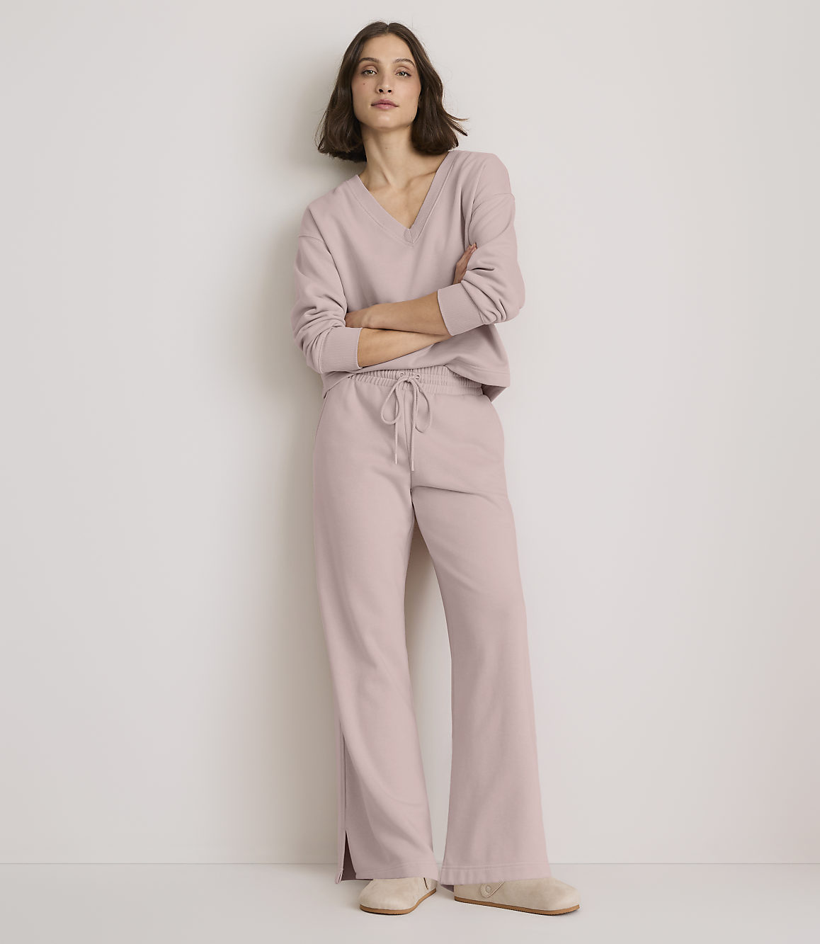 A woman in a light pink loungewear set stands with her arms crossed against a plain, light-colored wall.
