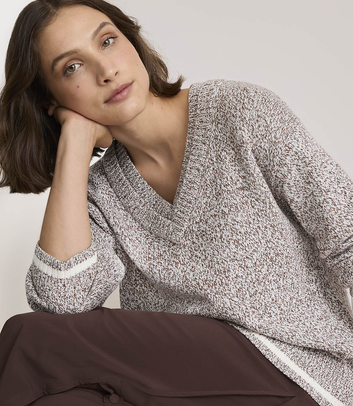 A woman in a speckled v-neck sweater rests her head on her hand against a plain background.