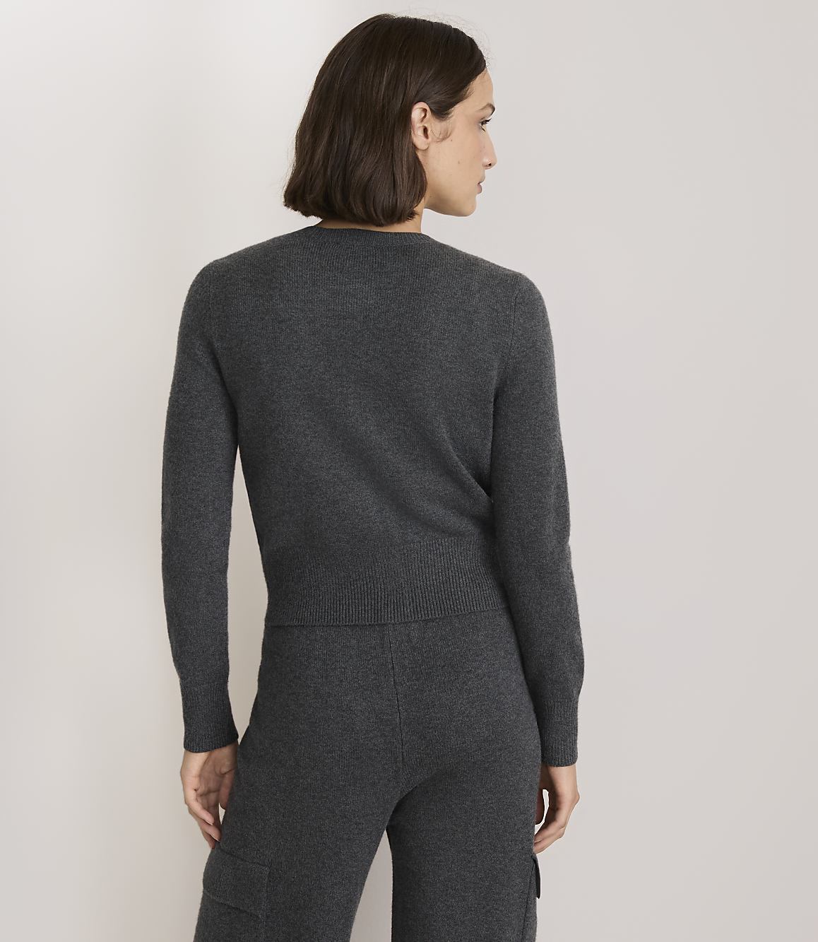A woman in a dark grey sweater and pants stands against a plain, light grey background.