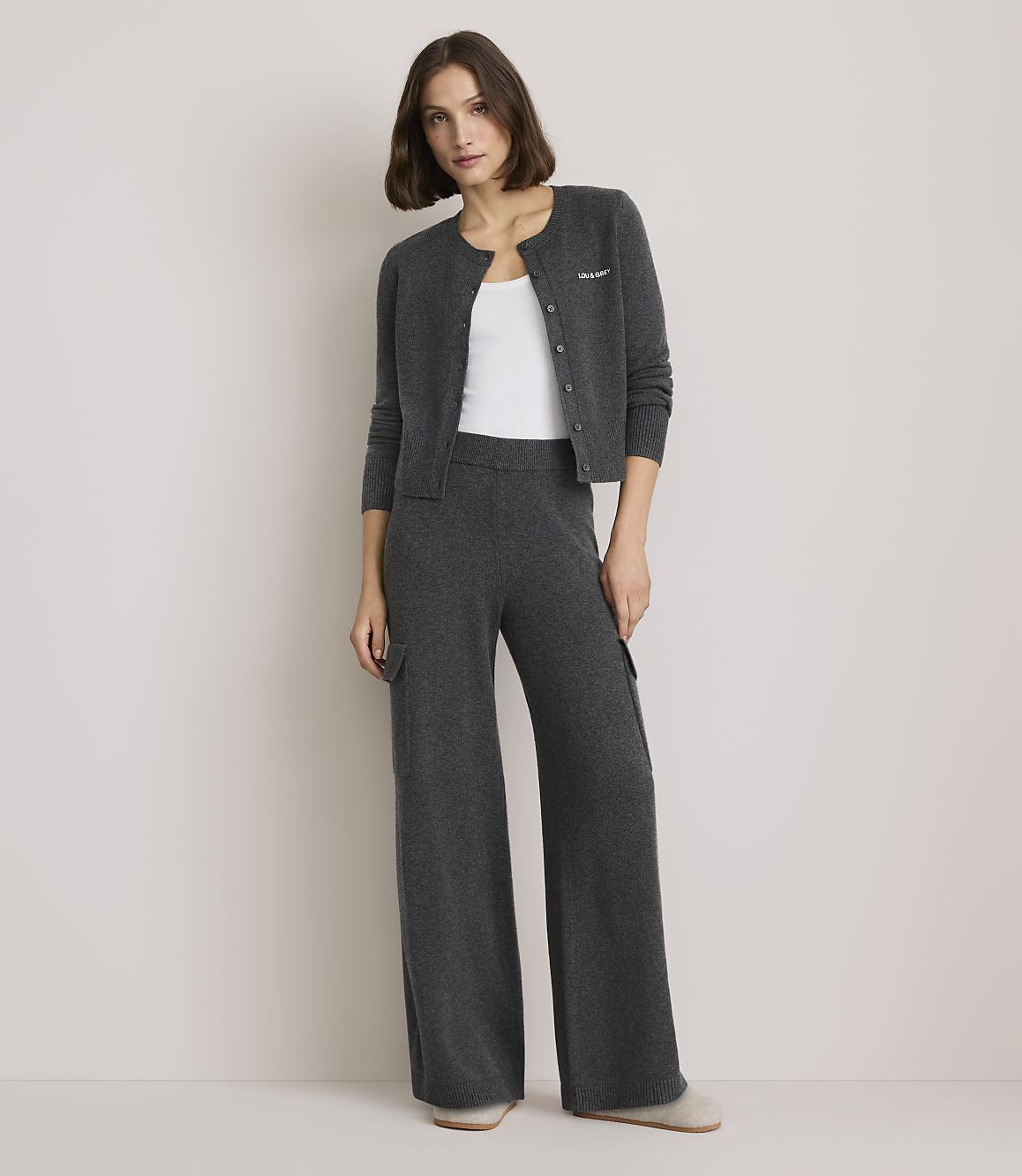 A woman in a grey cardigan and matching wide-leg pants stands against a plain, light grey background.