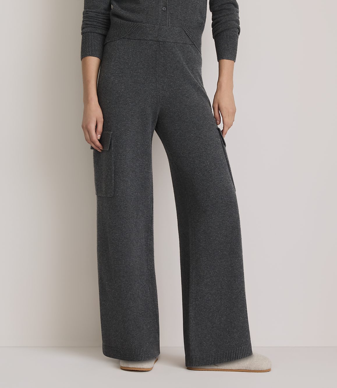 A person wearing dark gray wide-leg knit pants with cargo pockets stands on a light-colored floor, with a plain background.
