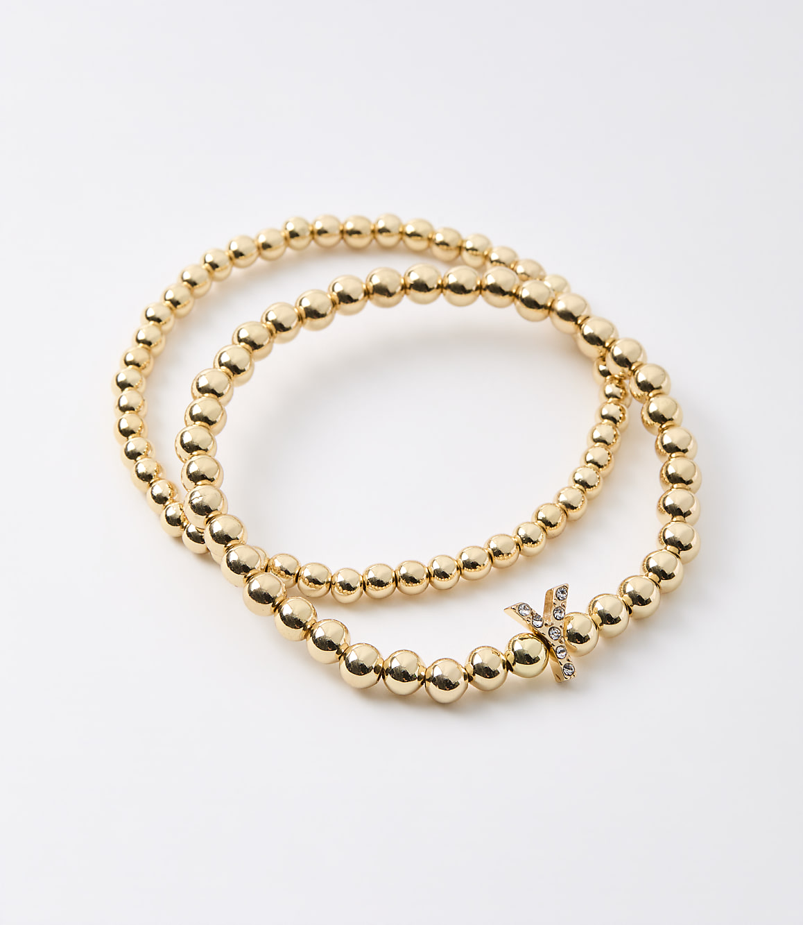 Two gold beaded bracelets, one with a diamond-studded "X" charm, are arranged on a plain white background.