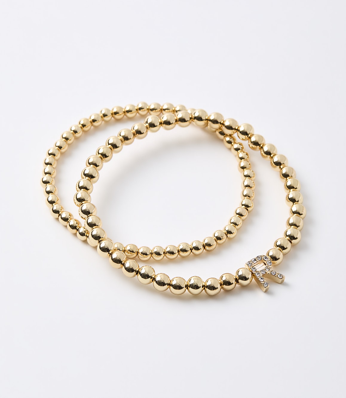 Two gold beaded bracelets, one with a diamond-encrusted "R" charm, are arranged on a plain white background.