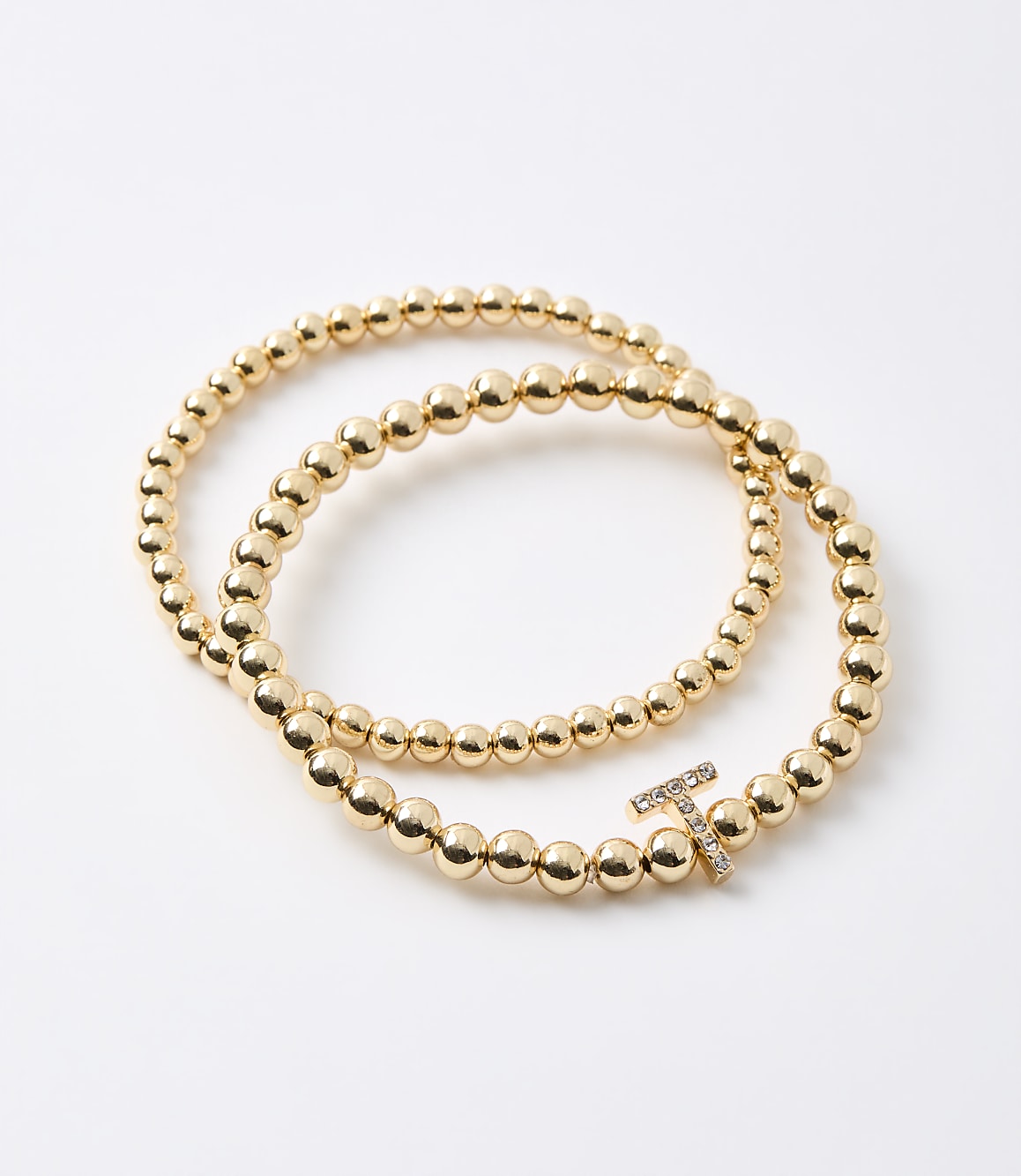 Two gold beaded bracelets, one with a diamond-encrusted "T" charm, lie on a plain white background.