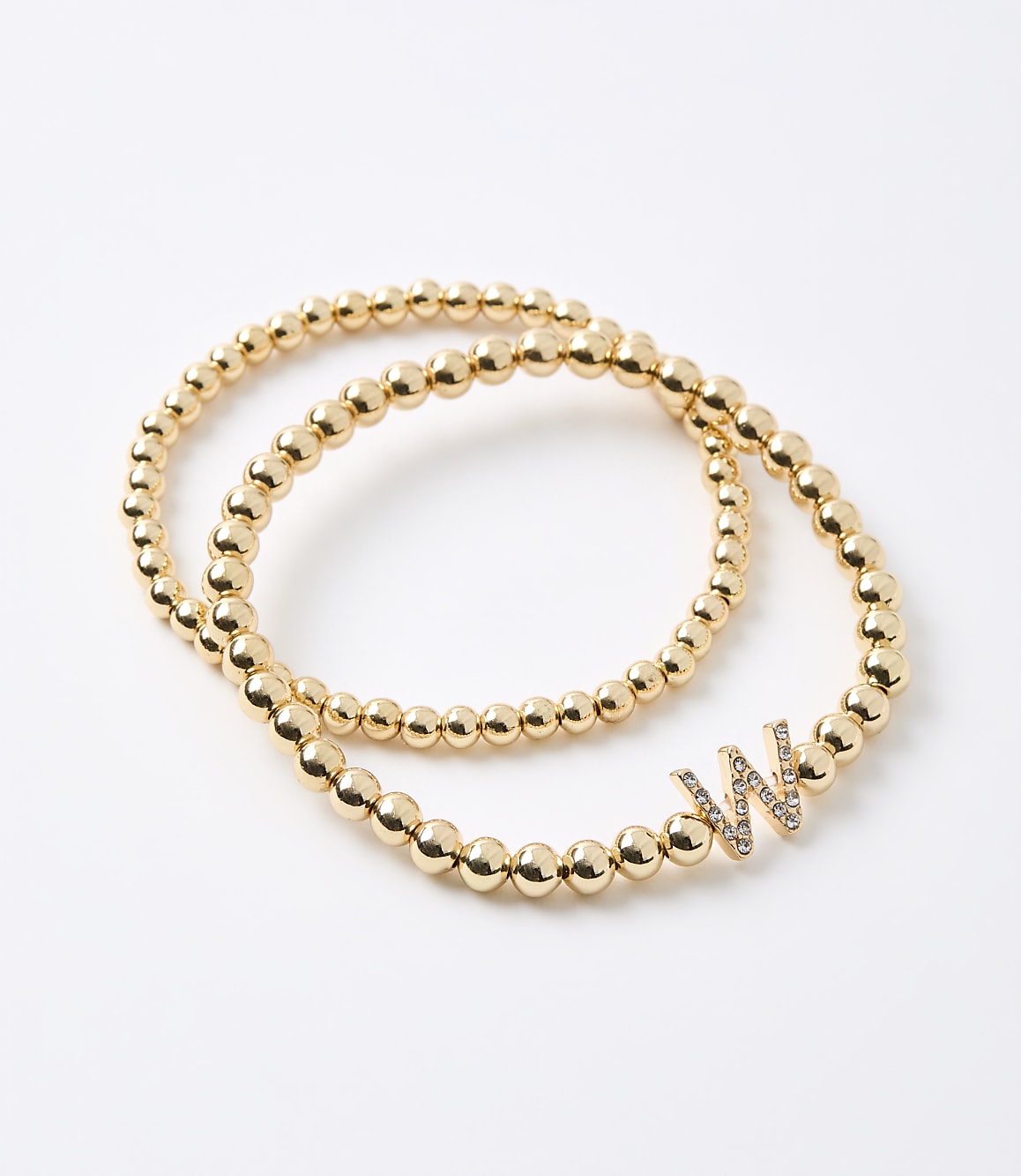 Two gold beaded bracelets, one with a diamond-encrusted "W" charm, rest on a plain white background.