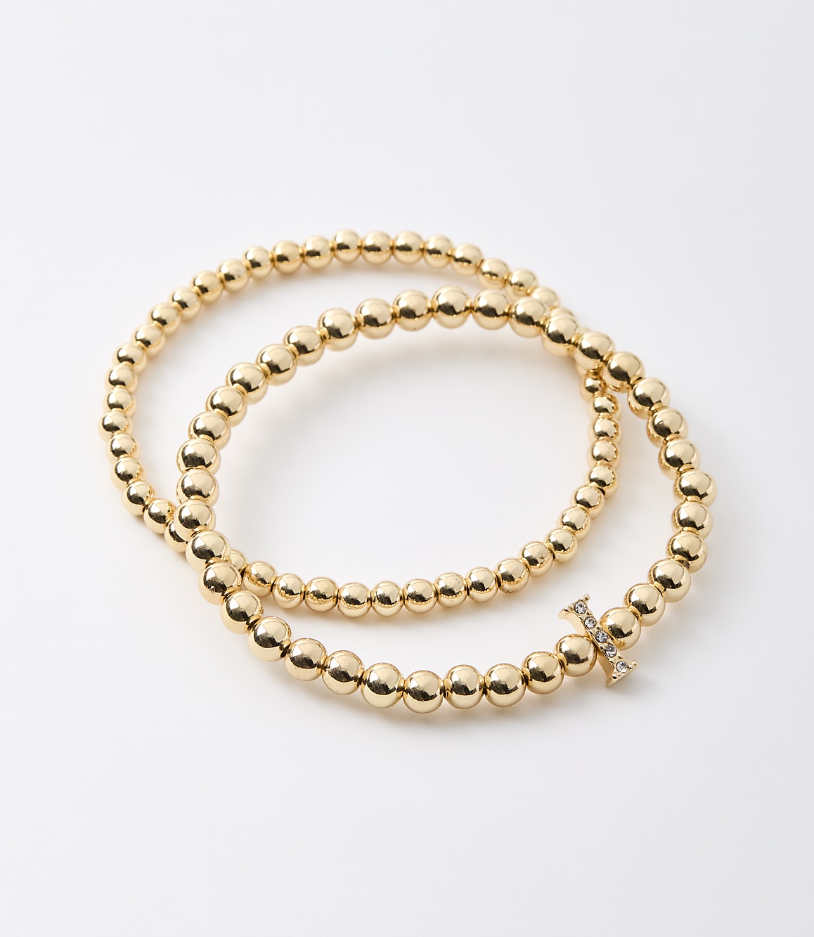 Two gold beaded bracelets, one with a diamond-encrusted "X" charm, are arranged on a plain white background.