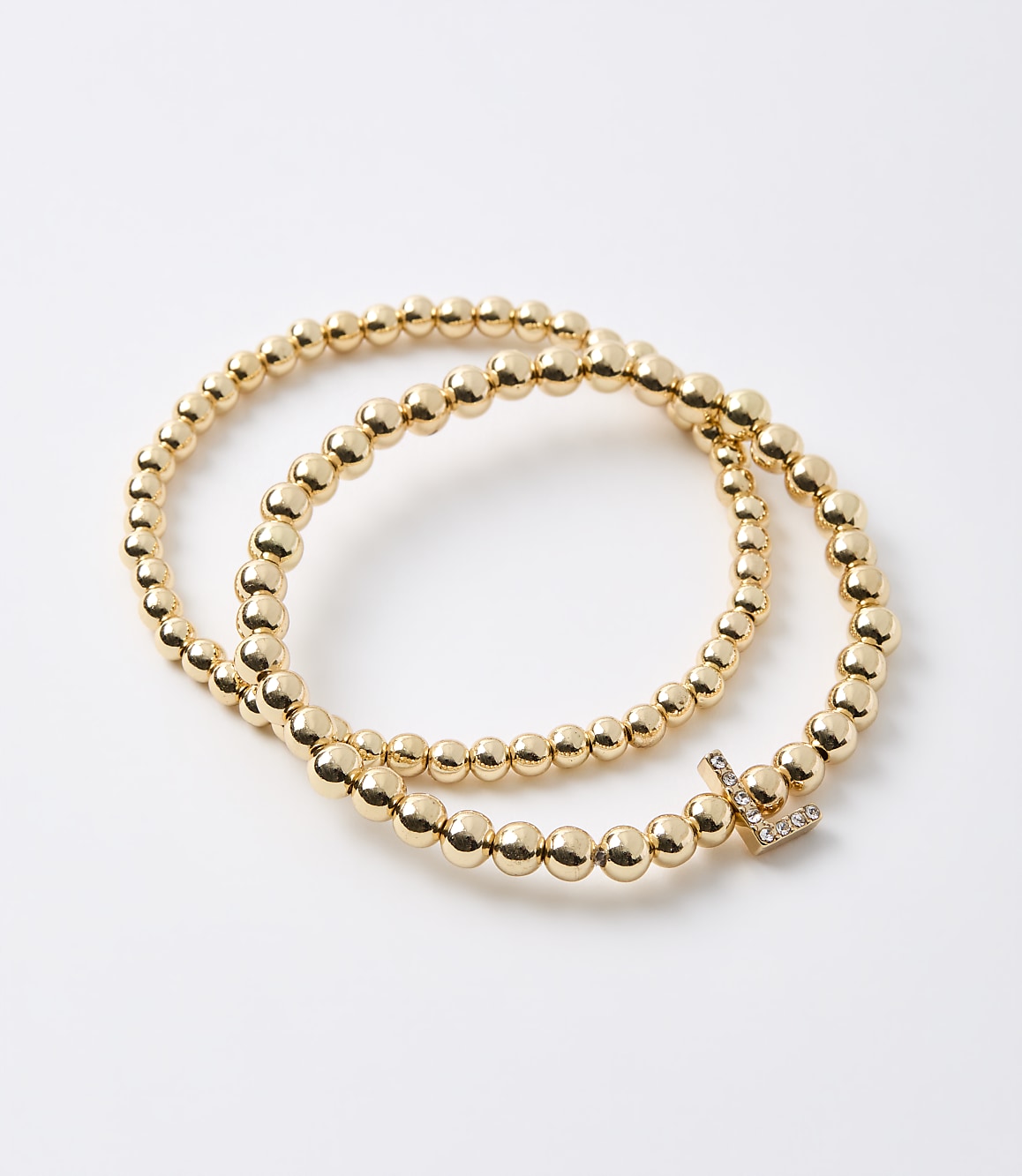 Two gold beaded bracelets, one with a diamond-encrusted initial "L" charm, rest on a plain white background.