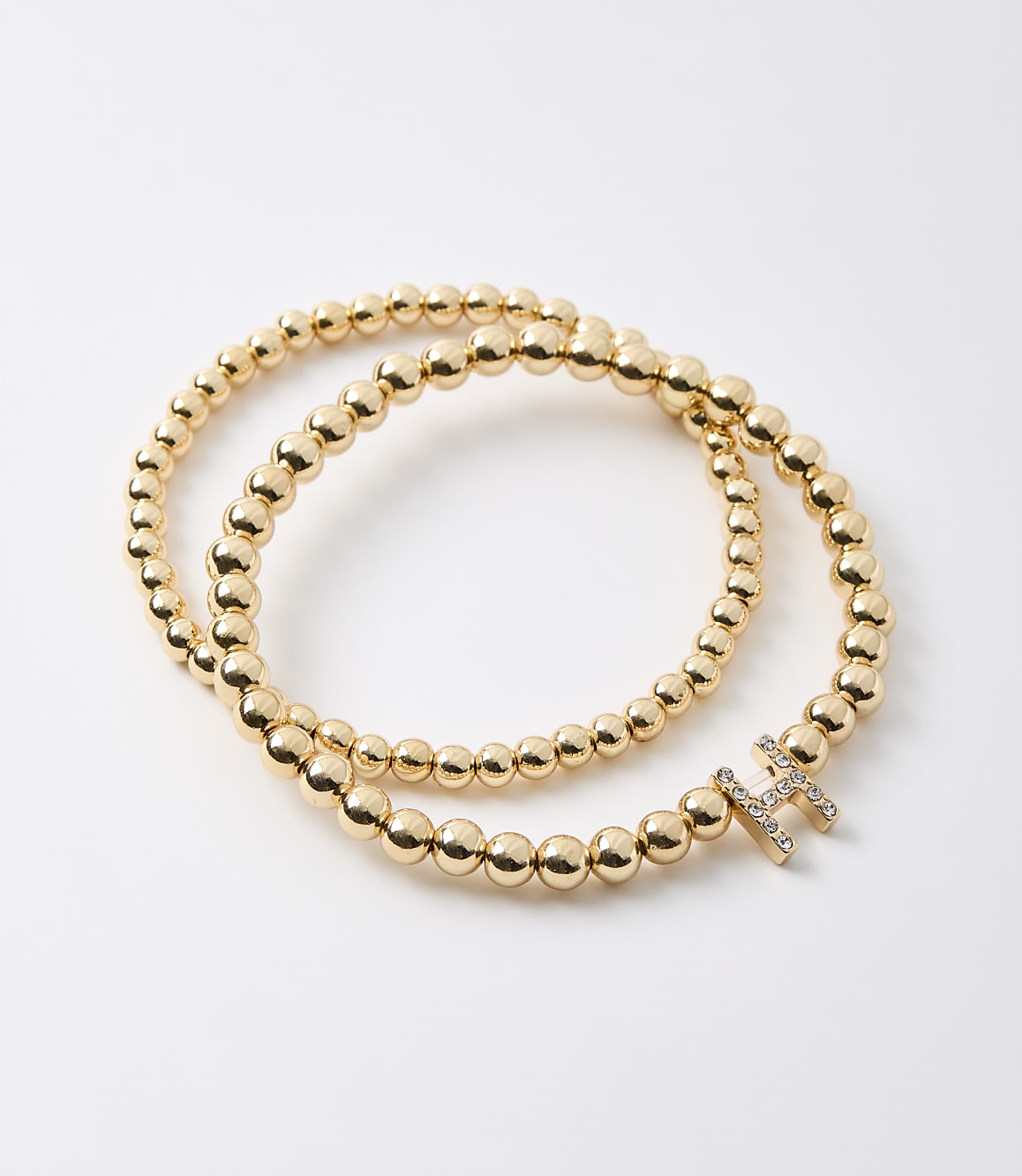 A gold beaded bracelet with a diamond-encrusted initial "H" rests on a plain white background.