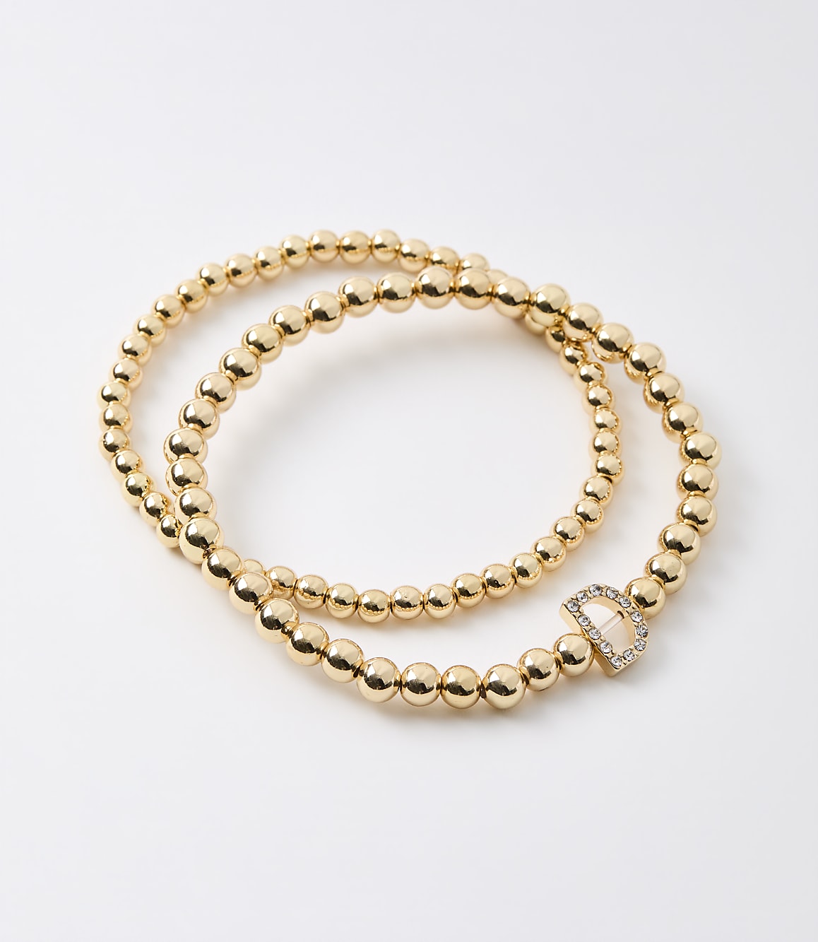Two gold beaded bracelets, one with a diamond-encrusted "D" charm, lie on a plain white background.