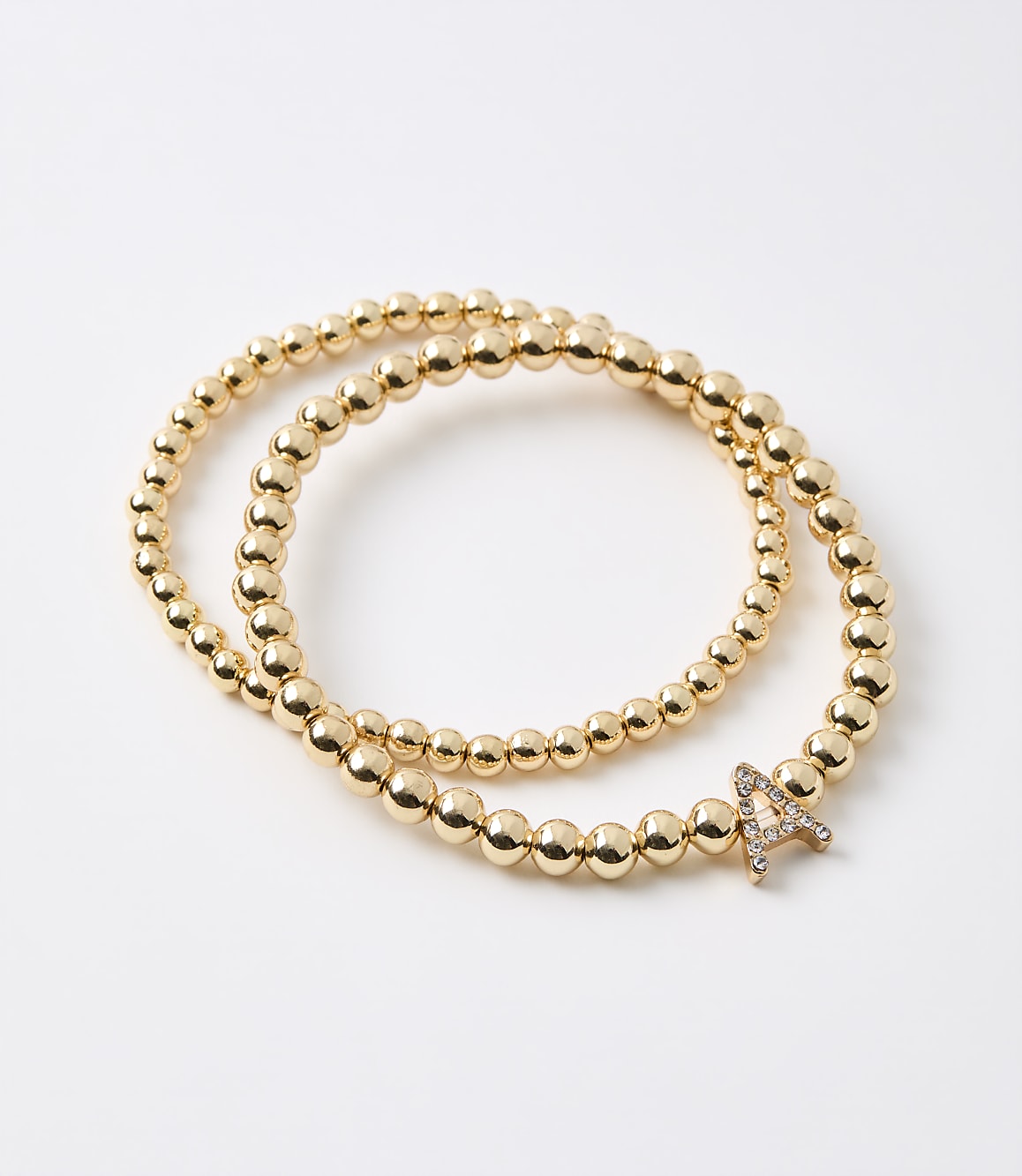 Two gold beaded bracelets, one with a diamond-encrusted letter "A" charm, lie on a plain white background.