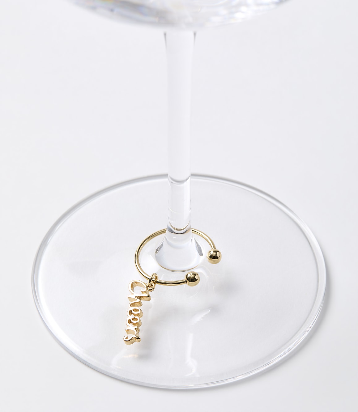 A gold "Cheers" charm hangs from a wine glass stem, set against a plain white background.