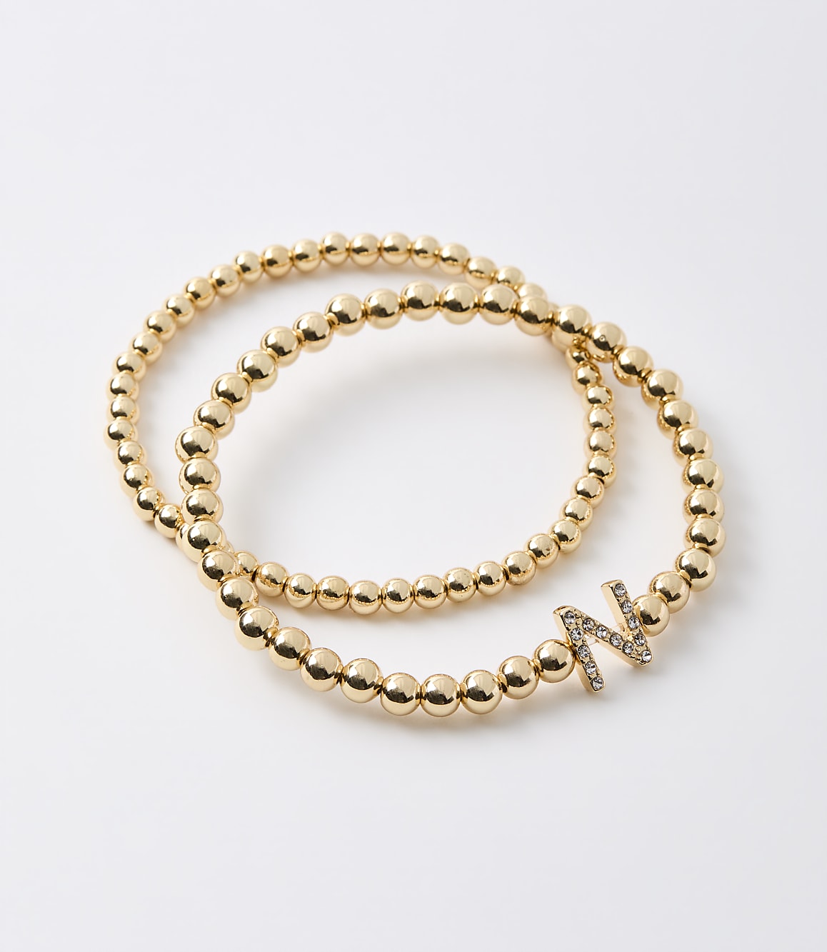 Two gold beaded bracelets, one with a diamond-encrusted letter "N" charm, lie on a plain white background.