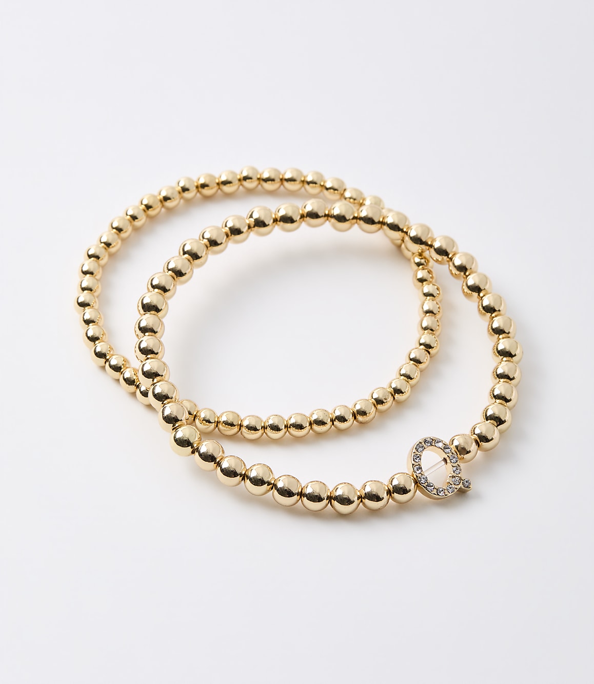 Two gold beaded bracelets, one with a diamond-encrusted "Q" charm, rest on a plain white background.