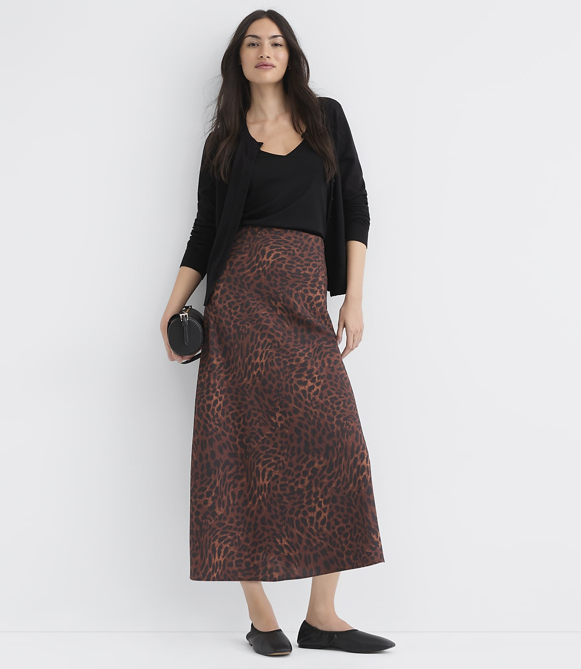 A woman in a leopard print skirt and black top stands against a plain white background, holding a small black purse.