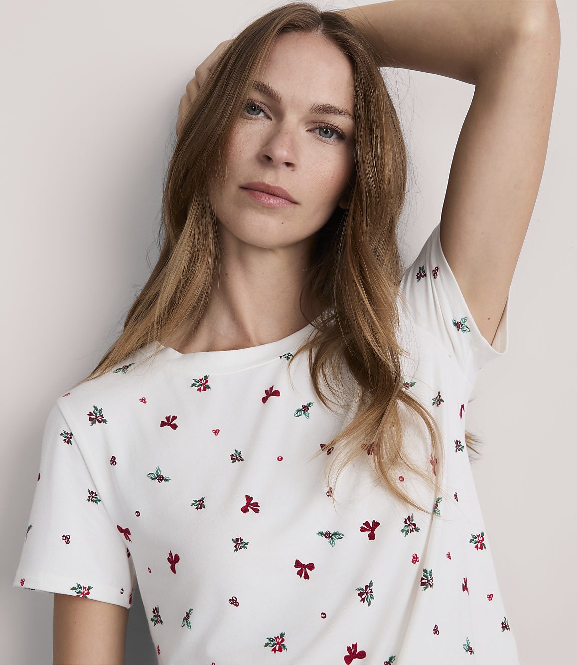 A woman with long, light brown hair and a white t-shirt with a subtle festive pattern poses against a plain, light-colored background.