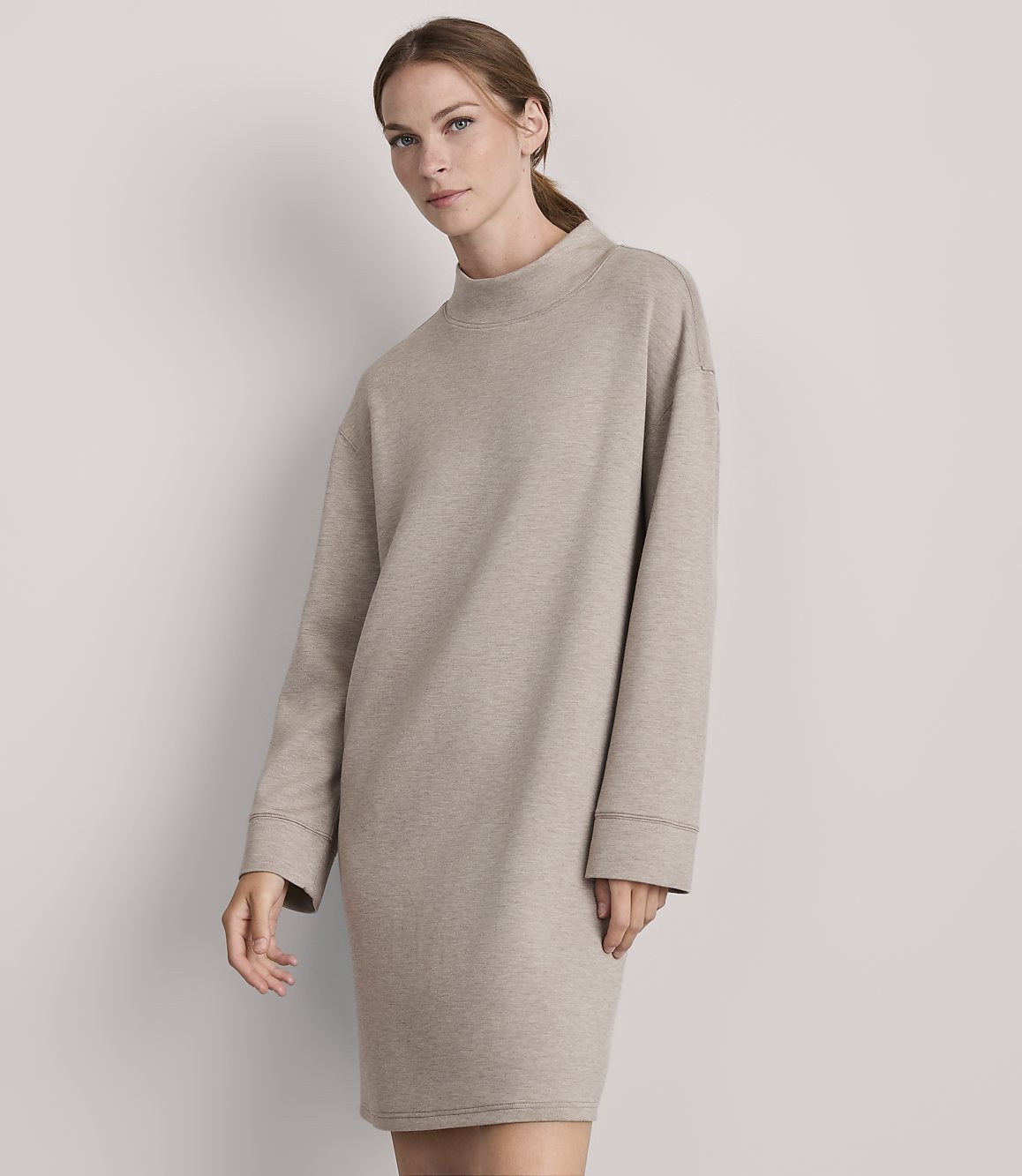 A woman in a beige sweatshirt dress stands against a plain, light gray background.