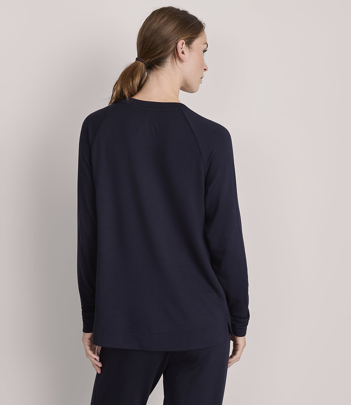 A woman in a dark blue sweater and pants stands with her back to the viewer against a plain, light gray background.