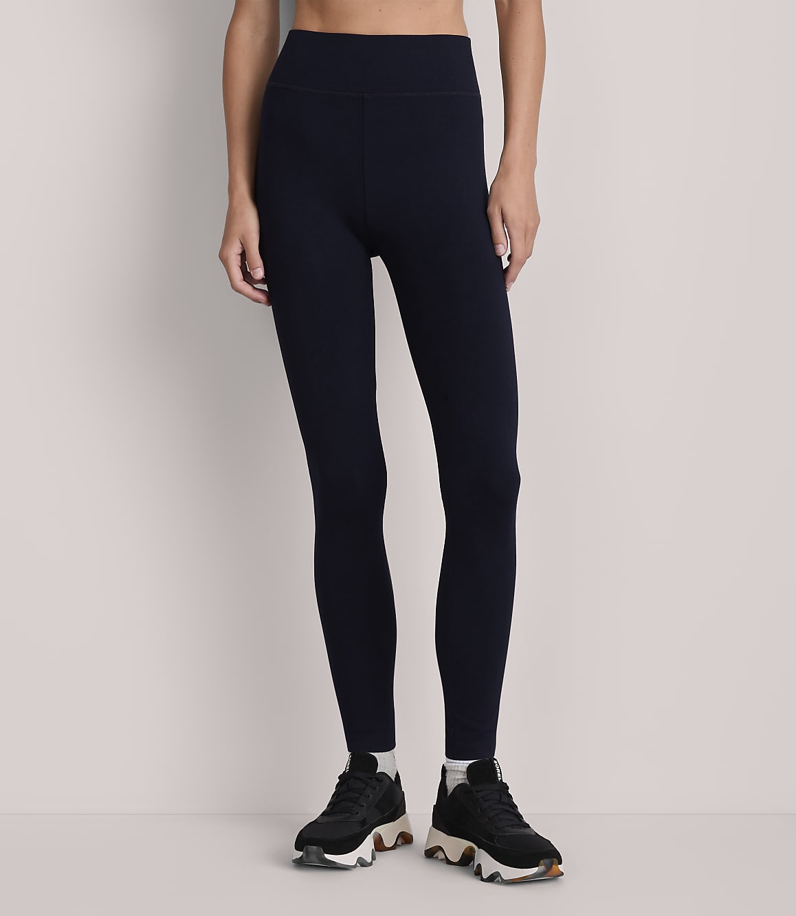 A person stands in dark blue leggings and black sneakers against a plain, light gray background.