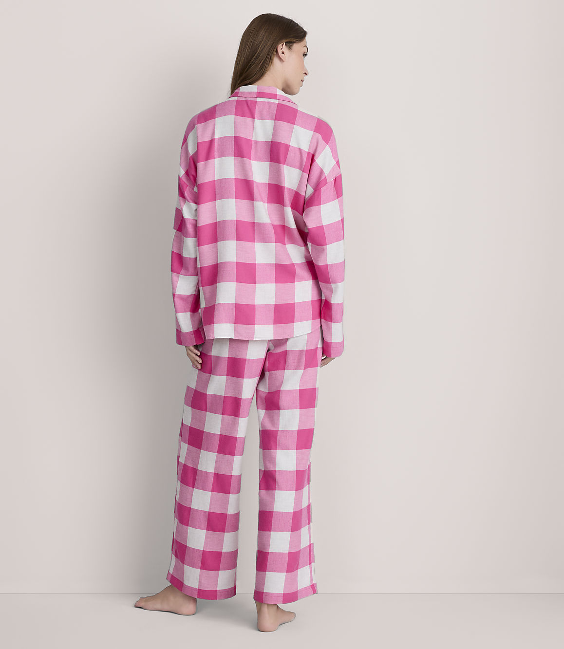 A woman stands facing away from the camera in pink and white checkered pajamas against a plain, light-colored background.
