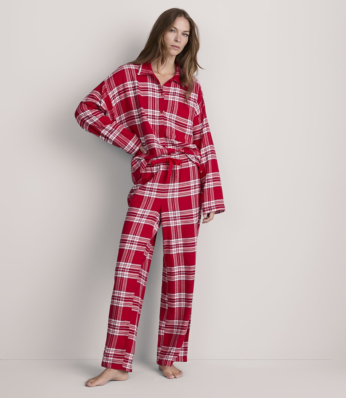A woman stands in red plaid pajamas against a plain, light-colored background.