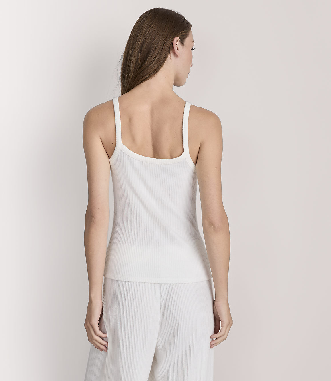 A woman in a white ribbed tank top and pants stands against a plain white background.