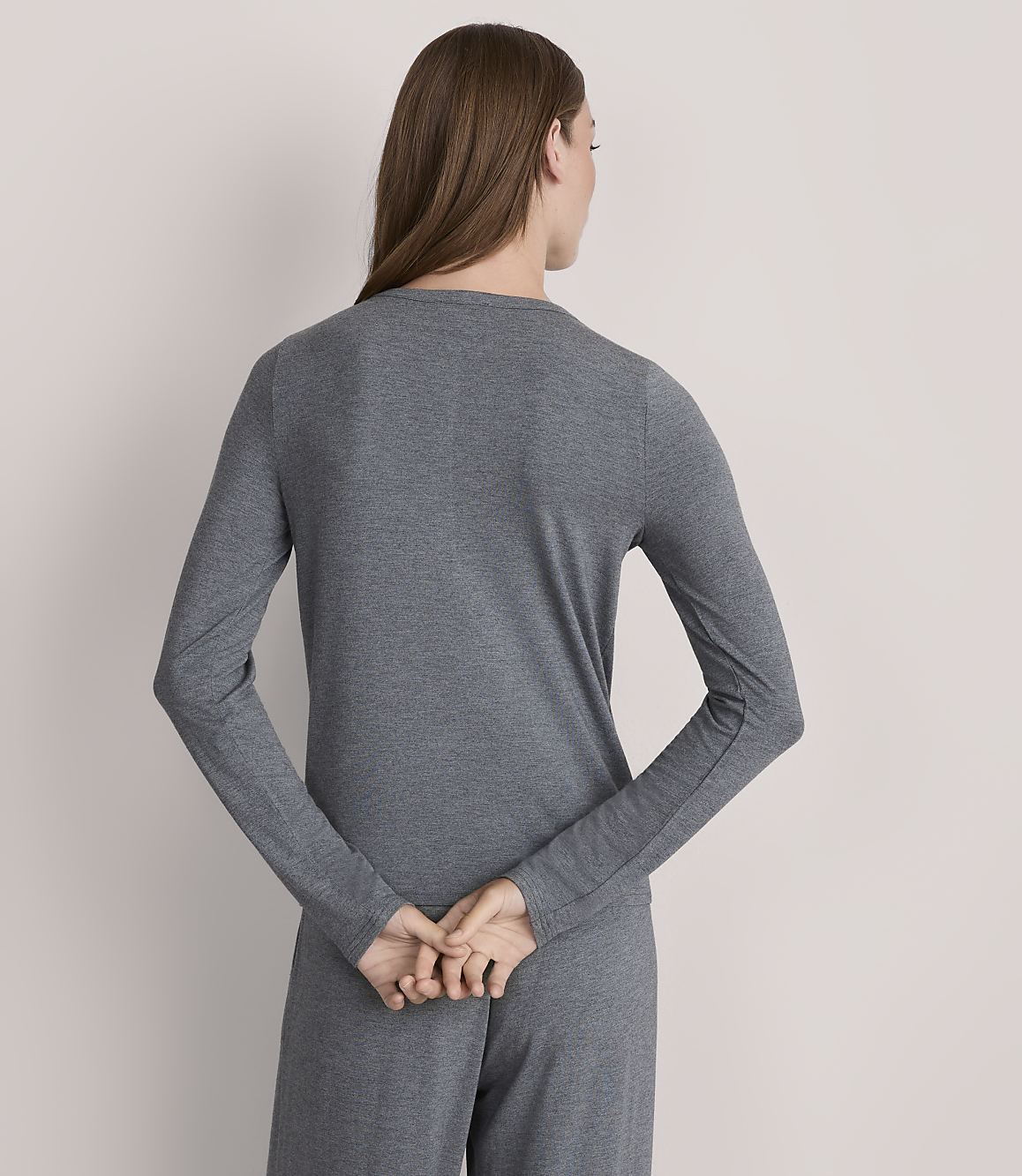 A woman in a grey long-sleeved shirt and pants stands with her back to the viewer against a plain grey background.