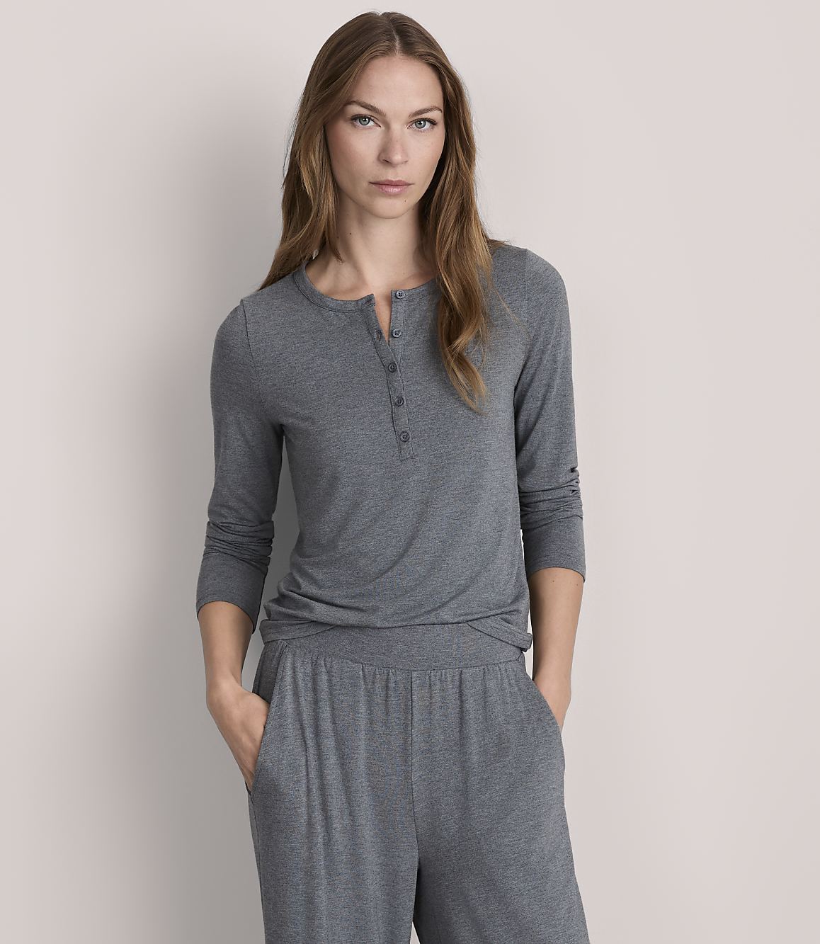 A woman in a grey henley shirt and matching pants stands against a plain, light grey background.
